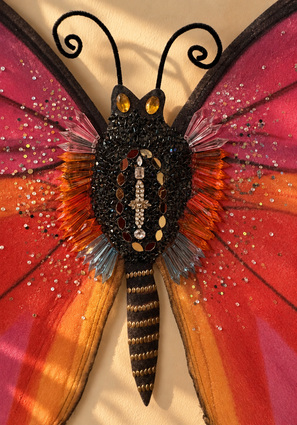 A highly detailed, colorful butterfly wall art piece featuring vibrant pink, orange, and red wings with silver glitter accents, a black textured body, and black antennae on a beige background.